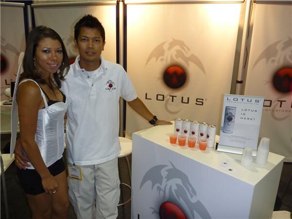 A man in a white polo shirt and a woman in a white corset top stand beside a counter holding Lotus cans and small orange sample cups, with Lotus dragon logos on the backdrop.