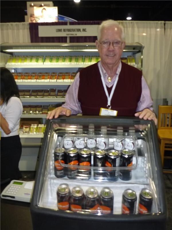 A smiling older man with glasses and a maroon vest stands behind a small open cooler of canned and bottled drinks, in front of a Lowe Refrigeration, Inc. display case.