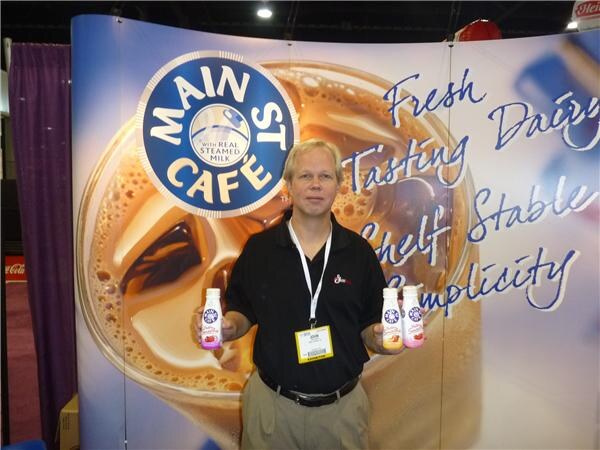 A man in a black polo shirt holds a small bottle in each hand in front of a large Main St Café backdrop showing iced coffee and the words Fresh Dairy Tasting, Shelf Stable.