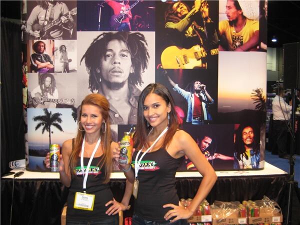 Two women in black Marley tank tops and badges hold Marley drink containers in front of a wall of black-and-white and color photographs of a musician. Cases of product sit below.