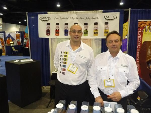 Two men in white MBS shirts and badges stand behind a table in front of an MBS banner showing bottles. A Grand Avenue Chocolates display is at the right edge.