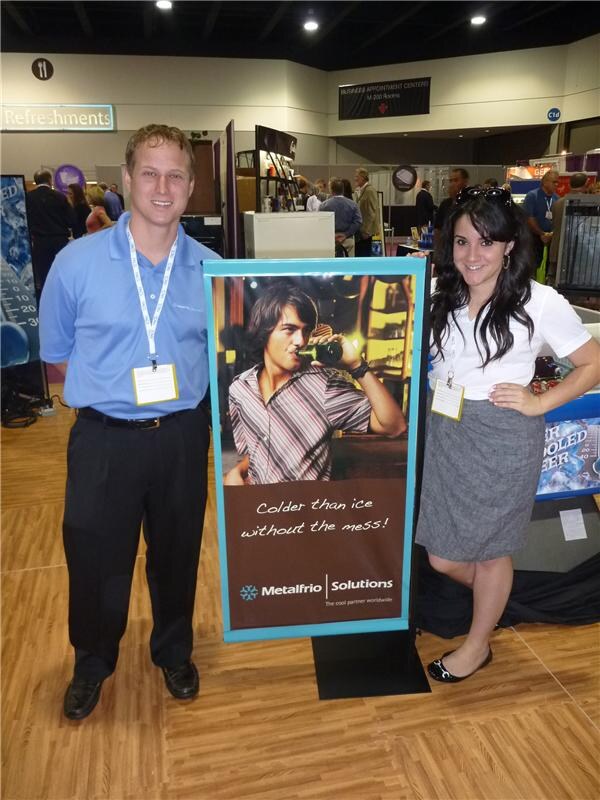 Two people stand on either side of a Metalfrio Solutions poster showing a man drinking from a bottle, with the text 'Colder than ice without the mess!' Both wear badges on a trade-show floor.