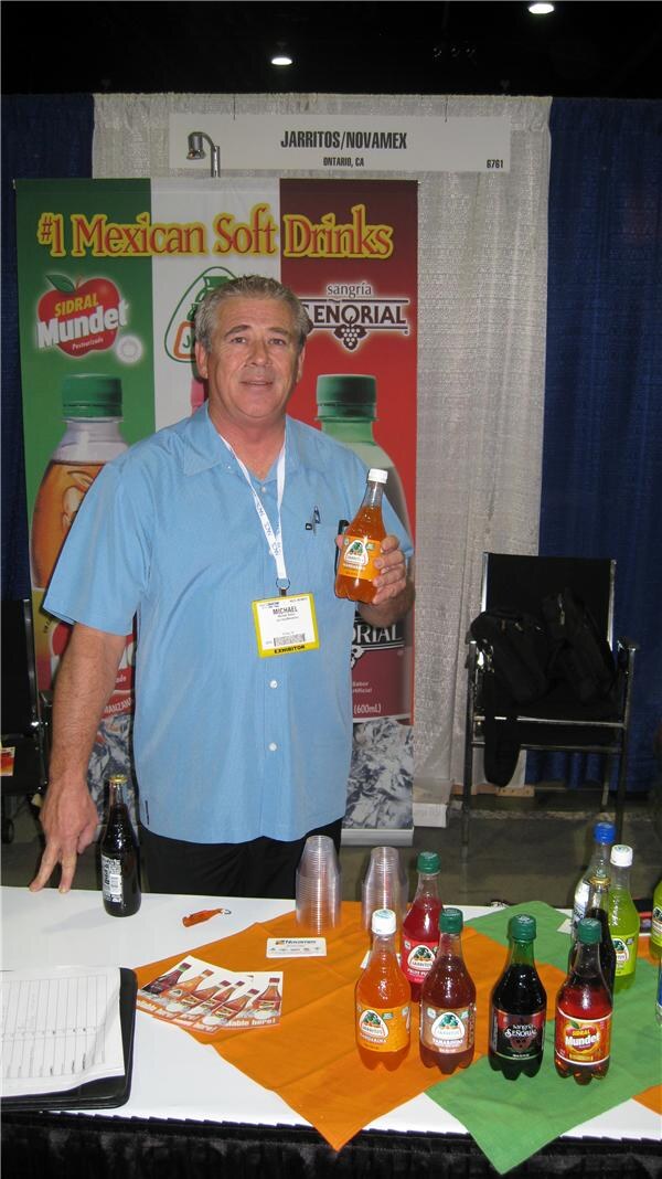 Michael, wearing a light blue shirt and badge, holds a bottle of Jarritos at the Jarritos/Novamex booth. Sidral Mundet and Sangría Señorial bottles sit on the table in front of him.