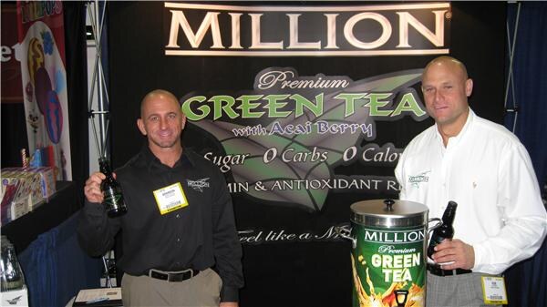 Two men stand in front of a black Million Premium Green Tea with Acai Berry backdrop, each holding a bottle. A Million Green Tea dispenser drum sits in front of one of them.