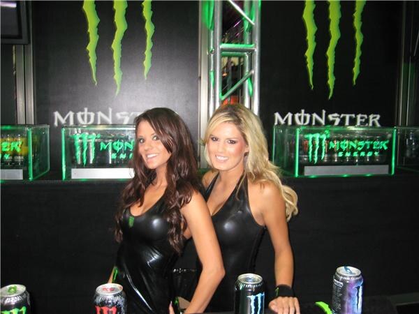 Two women in black outfits pose in front of Monster Energy signs with the claw logo, lit display cases behind them and Monster cans in the foreground.
