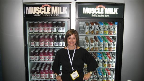 A smiling woman in a black top and lanyard stands with hands on hips between two Muscle Milk glass-door coolers stocked with bottled drinks.