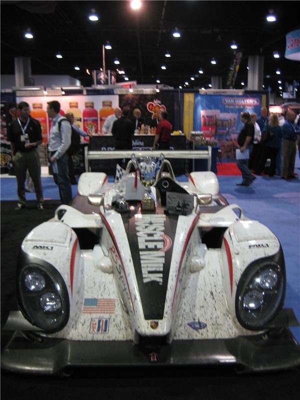 A white race car with Muscle Milk lettering on its nose is displayed on the show floor, with attendees and a Van Holten's booth with orange bottles in the background.