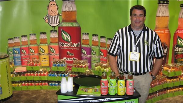 A man in a referee-style striped shirt stands at a green New Leaf booth with rows of New Leaf tea bottles, large bottle graphics and a cartoon referee figure behind him.