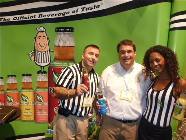 Two men and a woman pose in a New Leaf booth. One man and the woman wear referee-style striped outfits and hold bottles of tea. Bottles and a cartoon referee appear on the green backdrop.