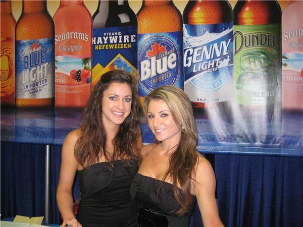 Two smiling women in black strapless tops stand in front of a banner of large beer bottles, including Labatt Blue, Labatt Blue Light, Genny Light, Dundee, Pyramid Haywire Hefeweizen and Seagram's Escapes.