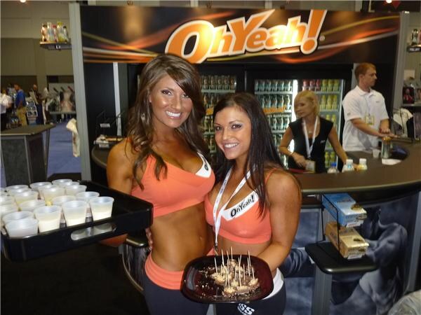 Two smiling women in orange athletic outfits hold a tray of small cups and a plate of snack samples at the OhYeah! booth, with refrigerated coolers and booth staff behind them.