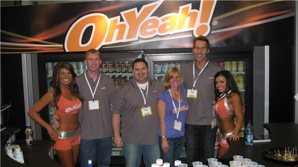 Three men and a woman wearing badges stand between two women in orange outfits in front of an OhYeah! sign and refrigerated coolers.