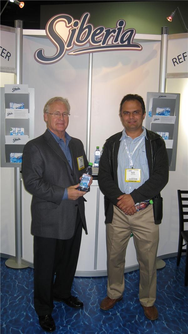 Two men stand in front of a Siberia booth. The man on the left holds a Siberia water bottle, and the man on the right wears a badge reading Oleg. Brochure holders hang on the walls behind them.