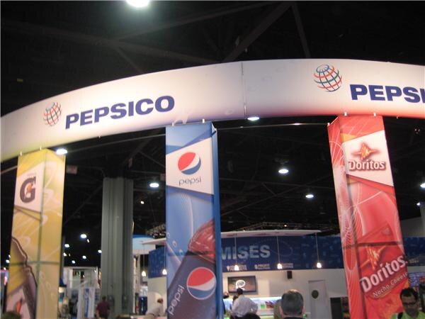 Overhead arch with PepsiCo signs above columns for Gatorade, Pepsi and Doritos, with visitors standing in the exhibit hall.
