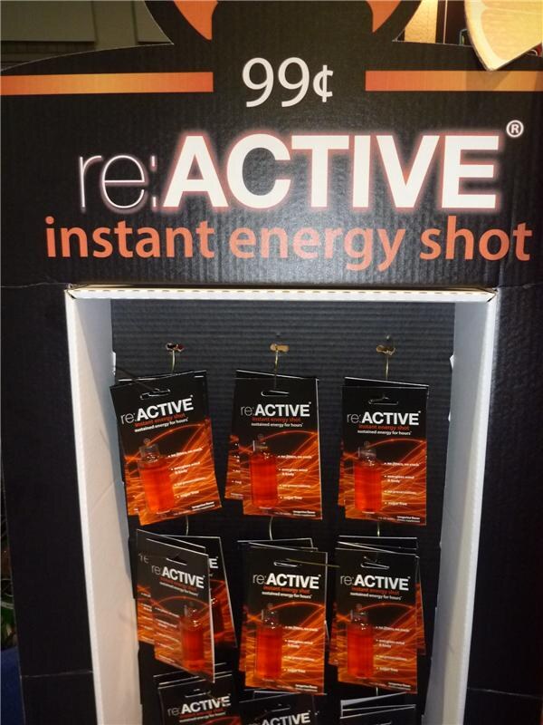 A cardboard display marked 99 cents and re:ACTIVE instant energy shot, with hanging packs of orange energy shot bottles in rows.