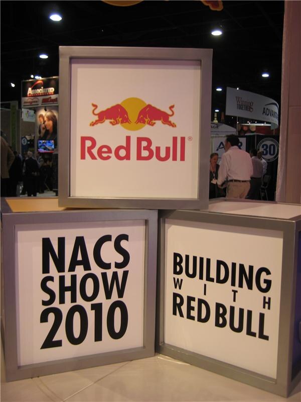 Stacked square display cubes: the top shows the Red Bull logo, the others read "NACS Show 2010" and "Building with Red Bull." Attendees and other booths are visible behind.