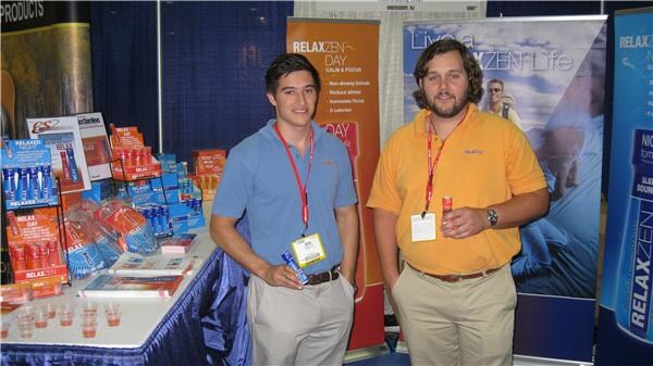 Two men wearing lanyards stand at a RelaxZen booth. Blue and orange RelaxZen product boxes sit on the table to the left, and banners behind them read Live a RelaxZen Life.