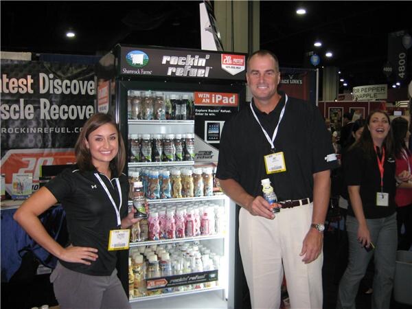 A man and a woman in black polos pose beside a Shamrock Farms Rockin' Refuel refrigerated cooler stocked with bottles. A woman stands smiling at right, and a Win an iPad sign is on the cooler.