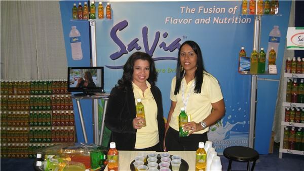 Two women in yellow polo shirts hold SaVia bottles at a booth with a blue backdrop reading The Fusion of Flavor and Nutrition. Cups and stacked bottles are displayed around them.