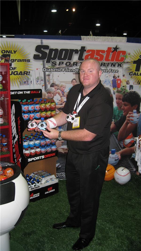 A bald man in a black polo holds two small ball-shaped Sportastic sports drink bottles in front of a Sportastic banner. Shelves of bottles and soccer and baseball props are around him.