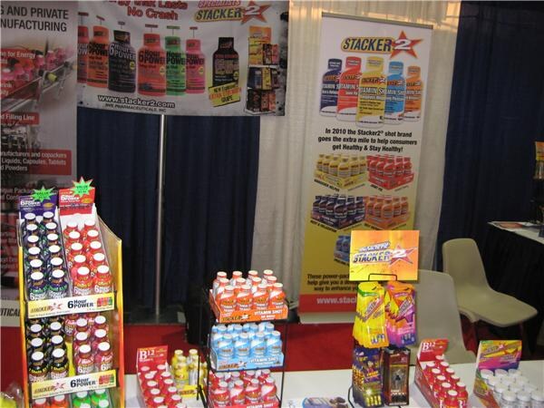 A booth display of Stacker 2 and 6 Hour Power shot bottles on racks and a table, with Stacker 2 banners behind them and an empty chair at the right.