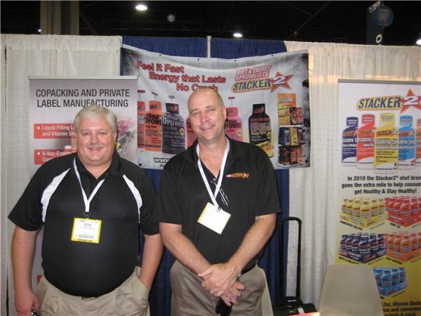 Two smiling men in black polo shirts and badges stand in a booth with Stacker 2 energy shot banners and a sign about copacking and private label manufacturing.