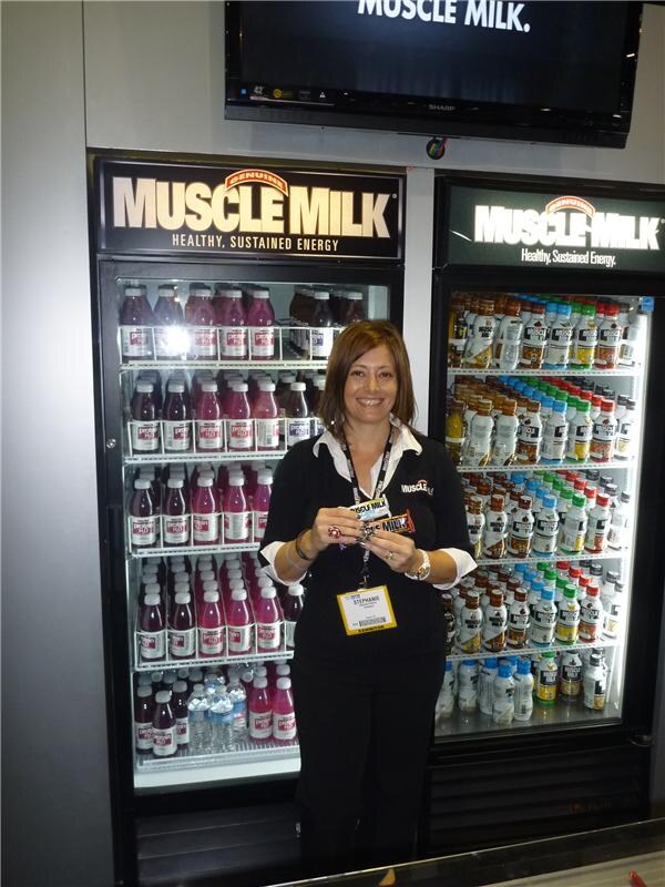 Stephanie, wearing a badge, stands holding two Muscle Milk bars between two lit Muscle Milk coolers filled with bottles. A Sharp monitor above shows the Muscle Milk name.