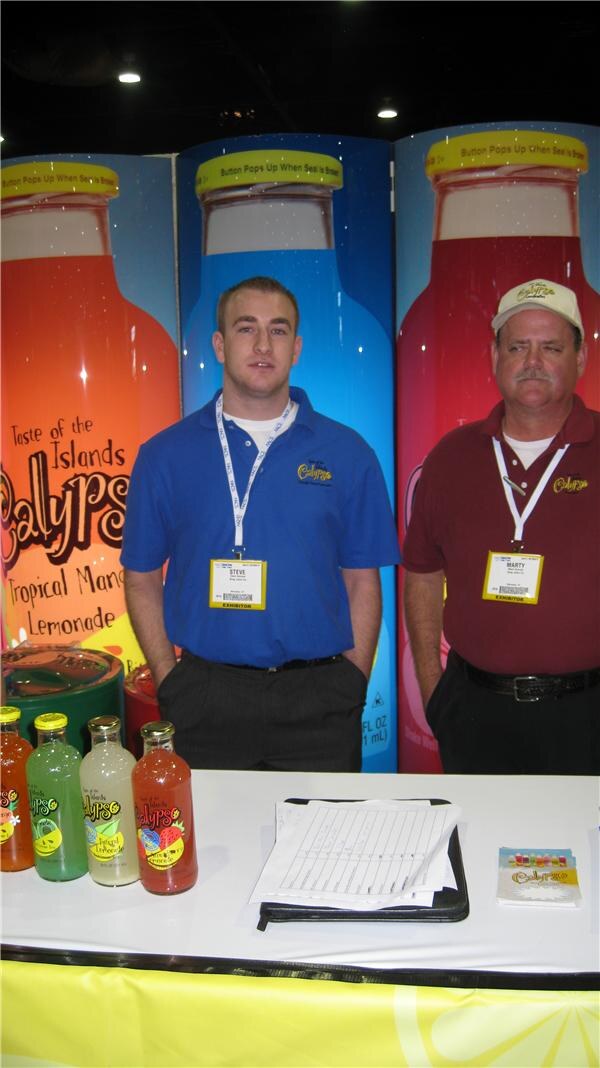 Steve, left, in a blue polo, and Marty, right, in a maroon polo and cap, stand at a table with Calypso lemonade bottles in front of large Calypso bottle graphics.