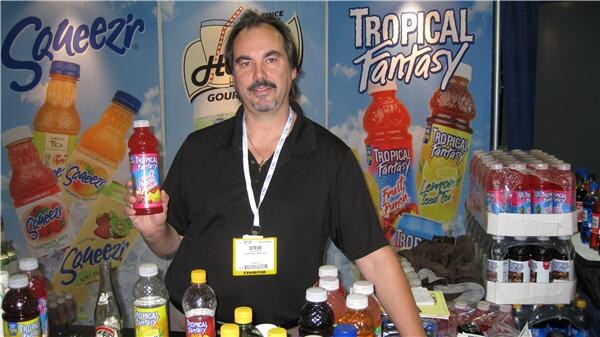 Steve, wearing a black shirt and an exhibitor badge, holds a bottle of red drink in front of Squeez'r and Tropical Fantasy banners, with bottled drinks on the table and shelf.
