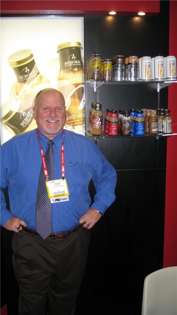 A smiling man in a blue shirt with an exhibitor badge reading Steve stands with hands on hips before Berner Food & Beverage iced coffee drinks on shelves.