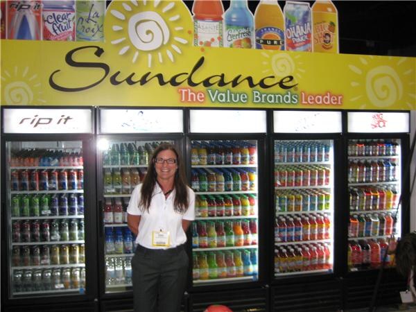 A woman in a white polo stands before five lit coolers under a yellow Sundance 'The Value Brands Leader' sign, with Rip It and La Croix among the beverages shown.