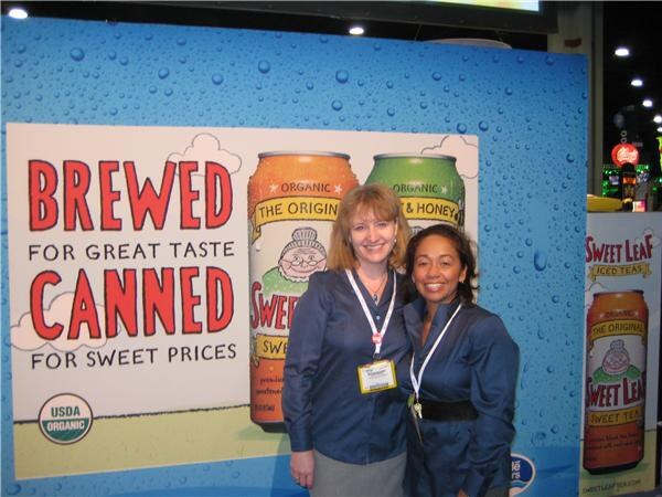 Two women in blue shirts and lanyards stand in front of a Sweet Leaf Tea banner showing canned iced tea and the words Brewed for great taste, canned for sweet prices.