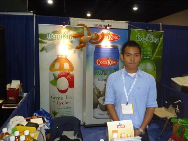 A man in a light blue shirt with a lanyard and badge stands in a booth with TropiKing Green Tea with Lychee and CocoKing Coconut Juice banners and a balloon monkey above.