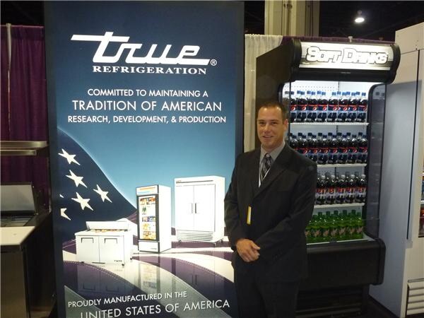 A man in a dark suit and tie stands beside a True Refrigeration banner and an open-air cooler labeled Soft Drinks, stocked with bottled soft drinks.