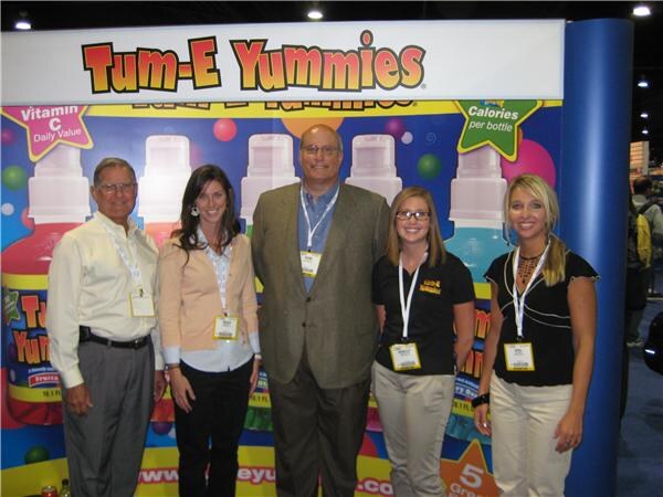Five people with badges, three women and two men, pose in front of a Tum-E Yummies display showing large bottle graphics.
