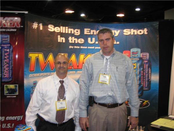 Two men with badges stand in front of a Tweaker banner reading #1 Selling Energy Shot in the U.S.A., with bottles of Tweaker shown on the backdrop.