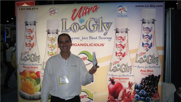 A smiling man in a white shirt holds a small bottle in front of a large Ultra Lo-Gly display showing Mango Mojito, Pomegranate and Acai-Blue juice bottles.