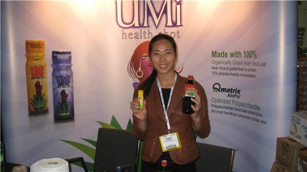 A smiling woman with a badge holds a small bottle and a larger bottle in front of a UMi health shot banner that mentions Qmatrix AloPol and aloe vera.