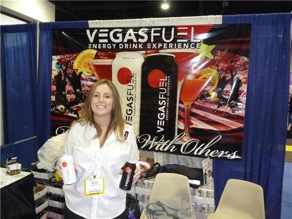A smiling woman with long hair and a name badge holds two Vegas Fuel energy drink cans in front of a Vegas Fuel Energy Drink Experience banner.