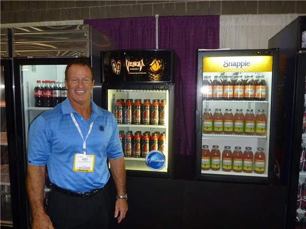 A smiling man in a blue polo and badge stands beside lit coolers stocked with Coca-Cola, Venom Energy and Snapple bottles.