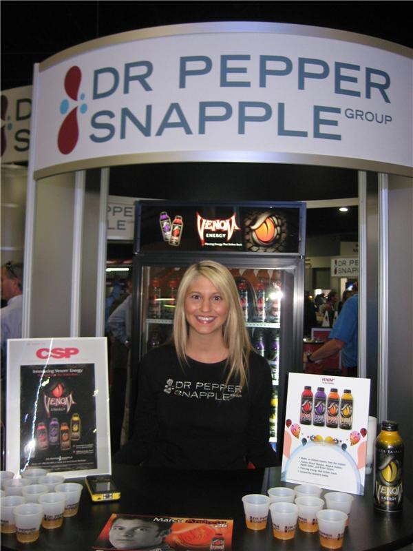 A smiling woman with blonde hair in a black Dr Pepper Snapple shirt stands behind a counter with sample cups, Venom Energy bottles and a lit cooler under a Dr Pepper Snapple Group sign.