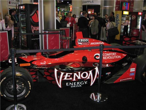 A red race car with Venom Energy Drink, Honda, Izod and Firestone logos sits behind stanchions at a Dr Pepper Snapple Group booth. A crowd stands in the background near a Venom cooler.