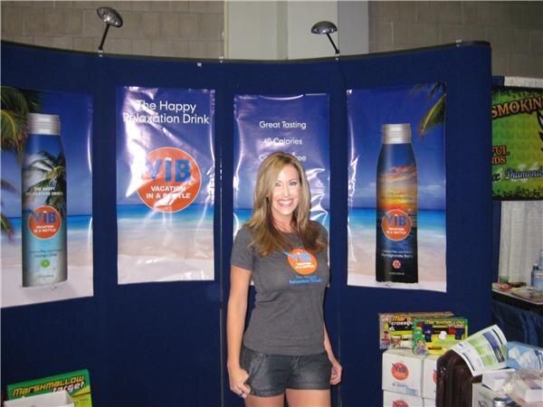 A smiling woman in a gray ViB t-shirt and denim shorts stands in front of blue ViB banners showing bottles on beach scenes and the words The Happy Relaxation Drink.