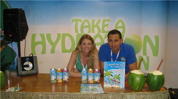 A woman and a man sit behind a table with Vita Coco cartons and two green coconuts with straws, in front of a Take a Hydration Vacation backdrop.