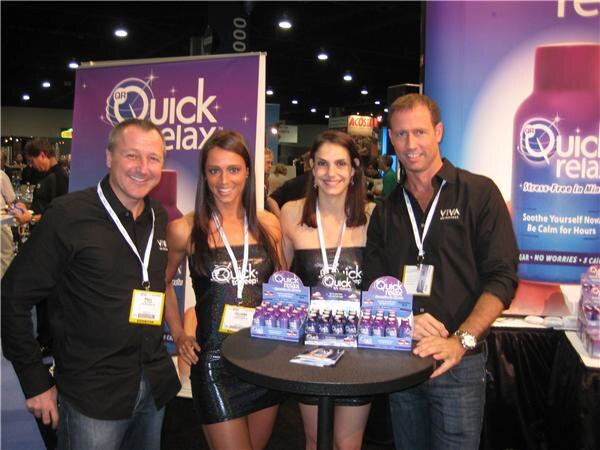 Two men in black Viva shirts and two women in black dresses stand around a table with Quick Relax display boxes of bottles, in front of Quick Relax banners.