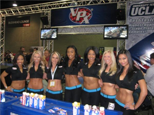 Seven women in black tops and blue-trimmed outfits stand in a row behind a table of VPX drinks, under a VPX sign with a UCLA banner nearby.