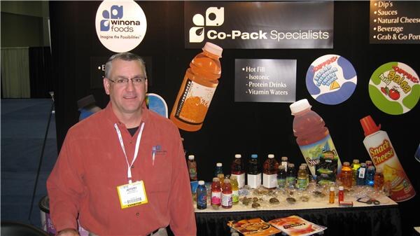 A smiling man in a red-orange shirt with a lanyard and badge stands in front of the Winona Foods booth, with Co-Pack Specialists signage and bottled drinks on a table behind him.