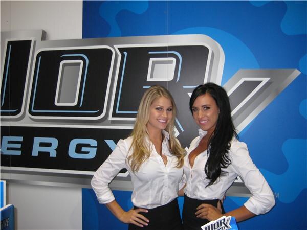 Two smiling women in white blouses and black skirts pose with hands on hips in front of a large Worx Energy logo on a blue wall.