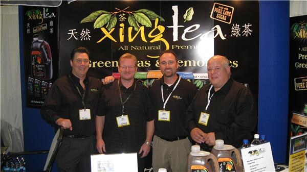 Four men with badges and lanyards stand side by side in front of a Xing Tea sign, with bottles of tea on the table in front of them.