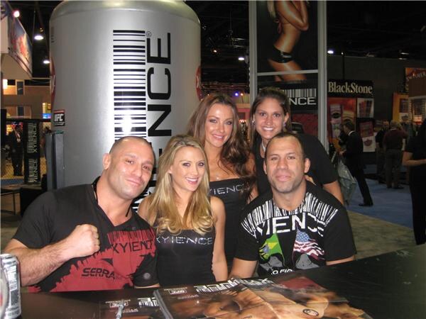 Two men and three women pose behind a table of magazines at the Xyience booth, with a large Xyience can display behind them and a BlackStone sign in the background.